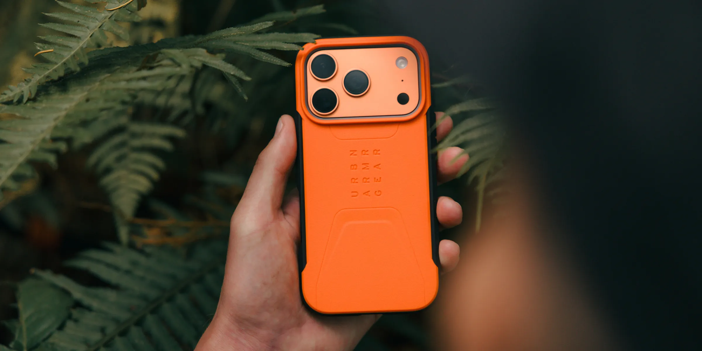 手握亮橘色 UAG Civilian 保護殼,背景是蕨類植物